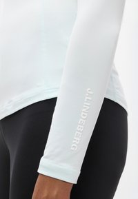 Ανοιχτό μπλε μπλουζάκι μακρυμάνικο από απαλό ύφασμα, με εφαρμοστή σχεδίαση και λευκό ανάγλυφο λογότυπο στο μανίκι. Ορατά μαύρα κολάν.