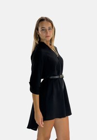 Robe noire avec un col et des manches longues, cintrée à la taille par une ceinture noire, avec un ourlet fluide.