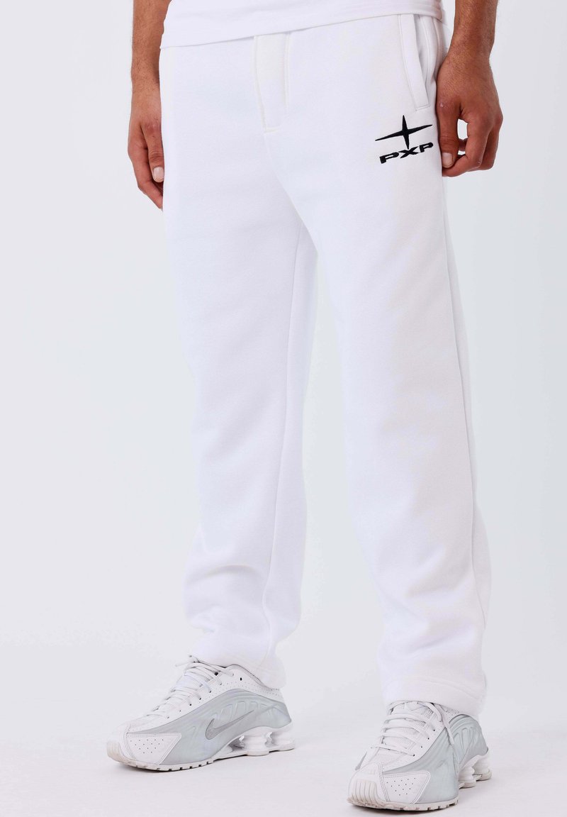 Man in witte joggingbroek met zwart "PXP" logo naast de zak, gecombineerd met witte en grijze Nike sneakers.