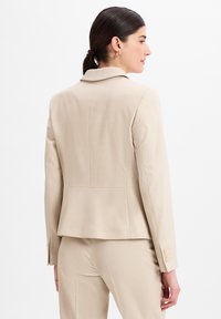 Blazer crème avec une coupe ajustée, en tissu texturé, col cranté et accents de trois boutons sur la manche. Porté sur un pantalon assorti.