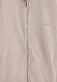Chaqueta de cremallera en color beige claro, fabricada con un tejido suave que presenta una textura lisa y un branding discreto. Incluye una lengüeta de tela en la cremallera.