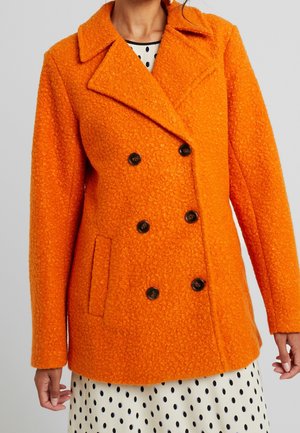 Kobieta ubrane w jaskrawo pomarańczowy, teksturowany, dwurzędowy płaszcz, na białej sukience w czarne kropki, stoi na jednolitym tle.