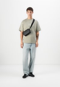 Ung mand iført en lysegrøn t-shirt med logo, lyseblå jeans, sorte sneakers og en sort crossover taske på hvid baggrund.
