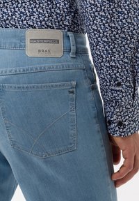 Acercamiento al bolsillo trasero de unos pantalones vaqueros de mezclilla azul claro con la etiqueta "Masterpiece BRAX", usado con una manga de camisa de patrón floral azul y blanco visible.