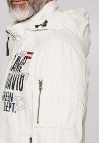 White hooded jacket with printed graphics, zippered pocket on the sleeve, and drawstring hood. Features a textured fabric.