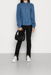 Mujer con blusa azul de botones, pantalones negros con dobladillo partido, zapatillas blancas, sosteniendo un pequeño bolso negro con asa trenzada.