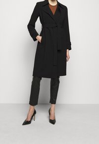 Schwarzer Trenchcoat mit einem taillierten Gürtel, Schalkragen, langen Ärmeln und Fronttaschen, kombiniert mit dunklen, enganliegenden Hosen und schwarzen Absätzen.