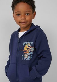 Marineblaues Zip-Hoodie aus Baumwollmaterial, mit buntem Tiger-Grafikdesign auf einem Fahrrad, zwei Fronttaschen, Rippstrickbündchen und -saum.
