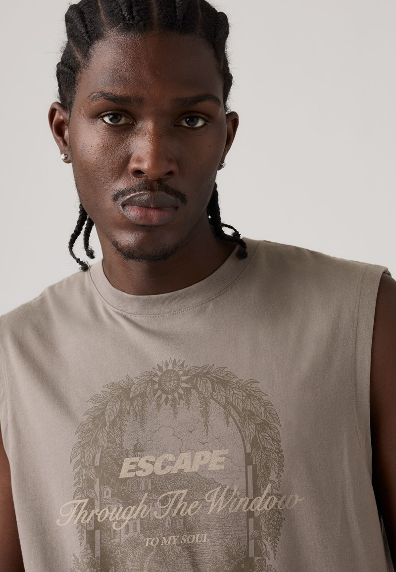 Junger Mann mit geflochtenem Haar, der ein ärmelloses beiges Hemd mit dem Schriftzug „ESCAPE Through The Window To My Soul“ und naturbezogenem Motiv trägt.