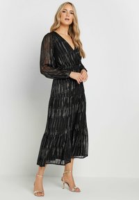 Svart maxi-klänning med V-ringning, genomskinliga långärmar och metalliska vertikala ränder. Har en lager-på-lager kjol med en flödande silhuett.