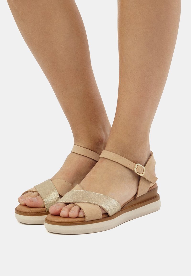 Beige sandaler med kryssremmer, en glitrende stropp, ankelrem med spenne, og en to-farget polstret såle. Jevn overflate og flat design.