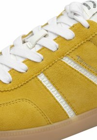Sneaker giallo in suede con lacci bianchi, design a strisce bianche e suola in gomma con finitura testurizzata. Branding sottile visibile.
