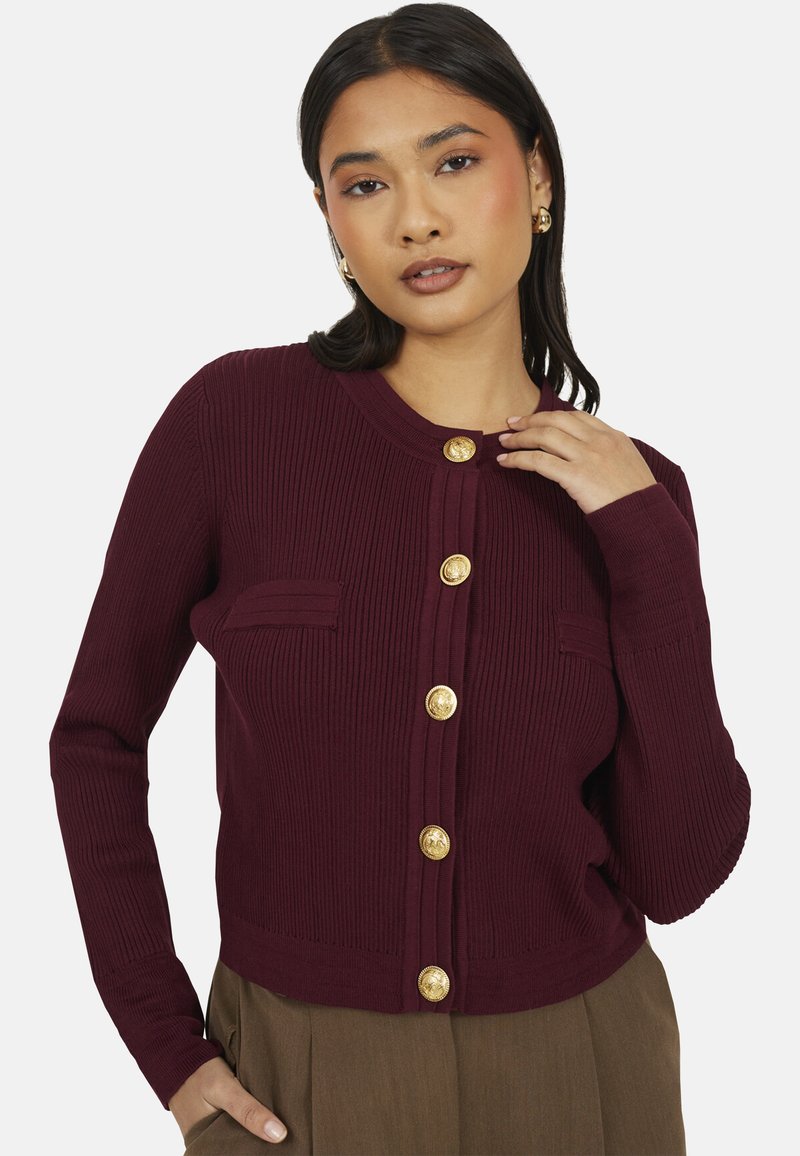 Cardigan côtelé bordeaux avec un col rond, mis en valeur par des boutons dorés et une poche poitrine unique. Tissu doux, design ajusté.