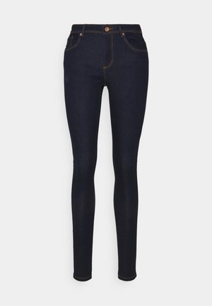 Donkere blauwe skinny jeans met vijf zakken, koperen klinknagels en contrasterende stiksels, gepresenteerd tegen een effen witte achtergrond.