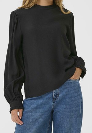 Kobieta ubrana w czarną bluzkę z długim rękawem z falbankami na mankietach oraz niebieskie dżinsy, ręka w kieszeni, na jednolitym tle.