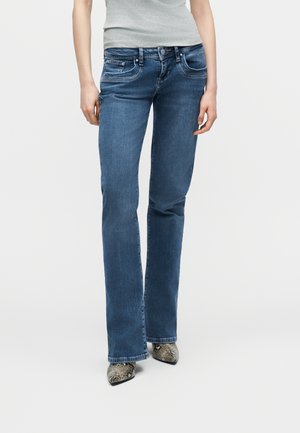 Nő közép-magas derekú kék bootcut farmert és szürke ujjatlan felsőt visel, hegyes kígyóbőr mintás cipőkkel, fehér háttér előtt áll.