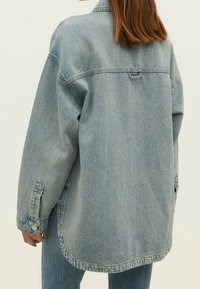 Persoon met schouderlang bruin haar, gekleed in een oversized lichtblauwe denimjacket en bijpassende jeans, met de rug naar de camera tegen een effen achtergrond.