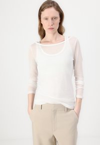 Langermet hvit topp laget av gjennomsiktig stoff, med rund hals og lagdelt design, sammen med lyse beige bukser.
