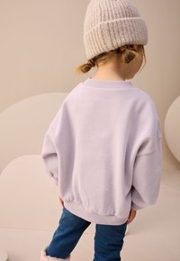 Lavandas krāsas pārmērīgi liels džemperis ar ribotu apkakli un aprocēm, kombinēts ar tumši zilām džinsa biksēm un gaiši bēšu adītu cepuri.