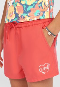 Korallfärgade badshorts med elastisk midja och dragsko, med en sidficka och en vit logotypdesign vid fållen. Slät textur.