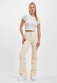 Ung kvinne som står, iført en hvit tettsittende kort T-skjorte, høyt liv beige bukser med knapper, og hvite sneakers mot en ensfarget bakgrunn.