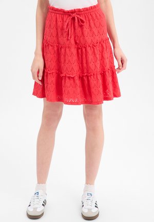 Vrouw draagt een rode gelaagde kanten rok met een taillekoord, witte sokken en witte Adidas sneakers met zwarte strepen.