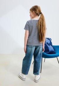 Paskowana koszulka w kolorze granatowym i białym, zestawiona z luźnymi niebieskimi dżinsami. Model ma na sobie białe wysokie trampki, stoi tyłem do widza.