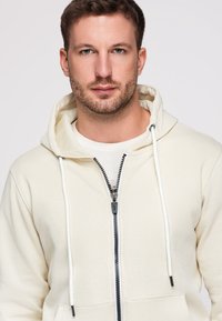 Jeune homme avec des cheveux courts marron et une barbe portant un sweat à capuche crème zippé sur une chemise blanche devant un fond clair uni.