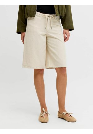 Kvinne iført beige knelang shorts med vertikal søm, knyttet midje, olivengrønn jakke og beige snøresko med flat såle.