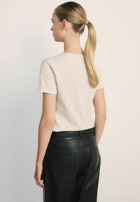 Kobieta z blondwłosym końskim ogonem, ubrana w beżową koszulę z krótkim rękawem i czarne skórzane spodnie z wysokim stanem, stojąca na jednolitym jasnym tle.