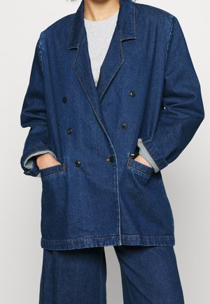 Osoba nosząca oversize'ową granatową dżinsową kurtkę dwurzędową na szarej koszuli, z rękami w kieszeniach, w połączeniu z dopasowanymi szerokimi dżinsowymi spodniami.