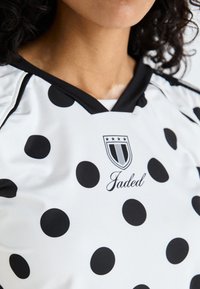 Bijela majica s velikim crnim točkama, s crnim V-izrezom i logom koji glasi "Jaded" s zvjezdicama, izrađena od glatkog materijala.