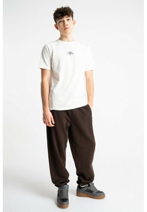 Młody mężczyzna o kręconych włosach stoi, ubrany w białą koszulkę z nadrukiem, luźne ciemnobrązowe spodnie i szare trampki na jednolitym białym tle.