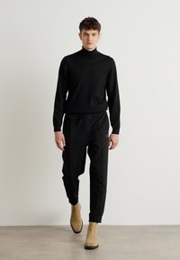Melnā turtlenecks jaqueta, apvienota ar brīva griezuma melnajām biksēm un brūniem potīšu zābakiem, veidojot monochromu izskatu ar kontrastējošu apavu akcentu.