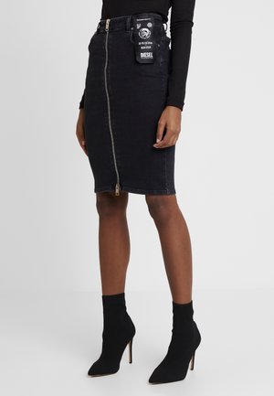 Frau trägt einen schwarzen knielangen Jeansrock mit Frontreißverschluss und Anhänger, kombiniert mit schwarzen Sock-Stiefeln und einem schwarzen Langarmshirt.