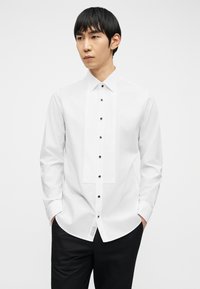 Νέος άνδρας με μαύρα μαλλιά που φοράει λευκό πουκάμισο με μαύρα κουμπιά και μαύρο παντελόνι, με τα χέρια στις τσέπες, κοιτάζει προς τα πλάγια.