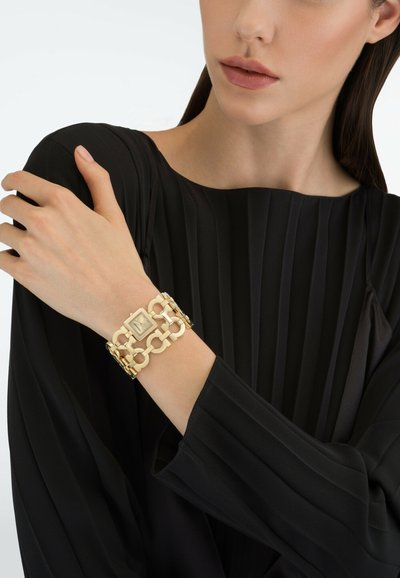 Mujer con una blusa negra plisada que lleva un reloj pulsera de eslabones dorados en la muñeca izquierda, con la mano descansando sobre su hombro derecho.
