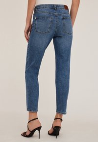 Kobieta w niebieskich, wysokich, prostych dżinsach oraz czarnych sandałach na wysokim obcasie, widoczna z tyłu.