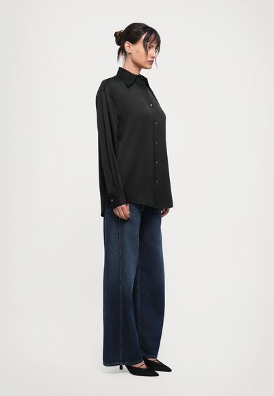 Schwarzes Langarmhemd mit Knopfschluss, kombiniert mit weit geschnittenen, dunklen Jeans und schwarzen spitzen Schuhen. Glatte Stoffbeschaffenheit.