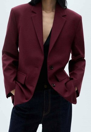 Personne portant un blazer rouge foncé avec deux boutons attachés, les mains dans les poches, associé à un pantalon foncé et un haut noir en dessous.