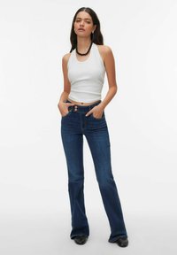 Witte haltertop gecombineerd met blauwe flared jeans met twee knopen aan de voorkant. Model staat met handen in de zakken en draagt een zwarte ketting.