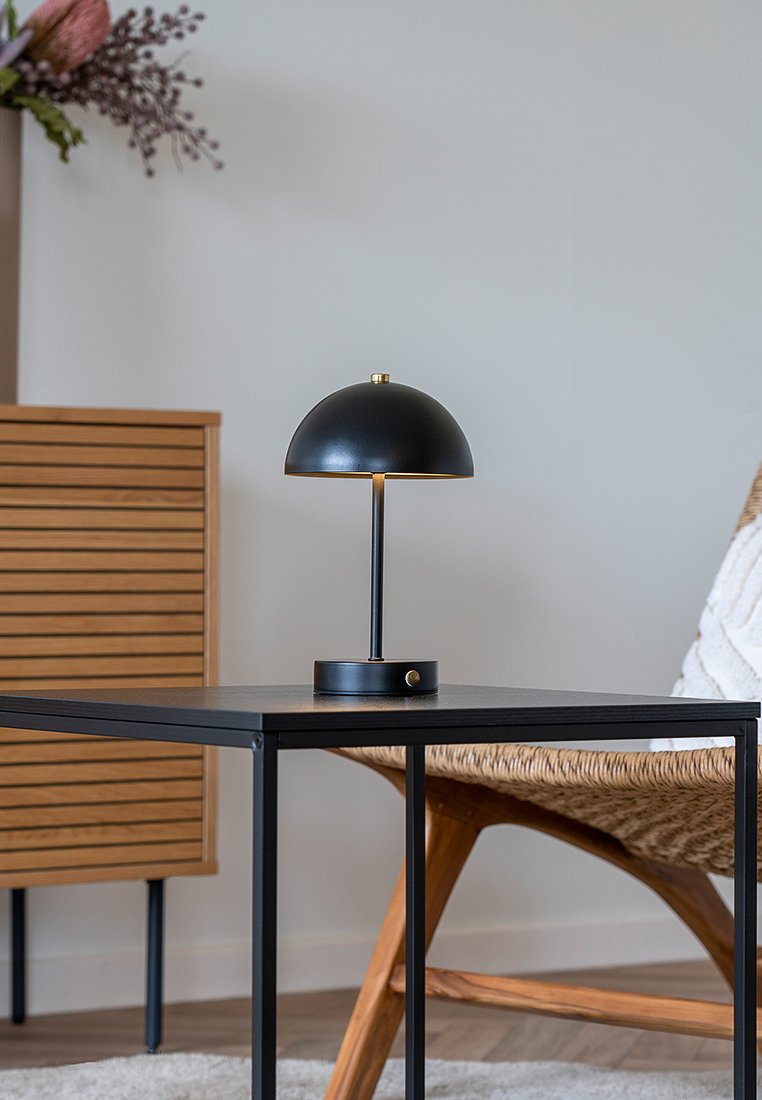 Lámpara de mesa de metal negra con pantalla en forma de cúpula y un acento de latón, situada sobre una mesa negra al lado de una silla de mimbre.