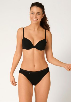 Kobieta o długich ciemnych włosach uśmiecha się, ubrana w czarny biustonosz i dopasowaną czarną bieliznę na jasnym, jednolitym tle.