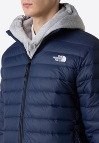 Giacca puffer blu navy con texture trapuntata verticale, cerniera frontale e accento di logo bianco; indossata sopra una felpa grigia con cappuccio.