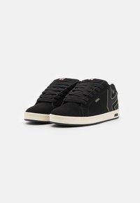 Black suede sneakers with a white rubber sole, featuring black laces and a contrasting green logo on the side. Textured upper with stitching details.