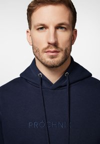 Granatowy bluza z kapturem z dekoltem w serek, sznurkiem do regulacji i haftowanym logo. Gładka tekstura z żeberkowymi akcentami przy dekolcie i rękawach.