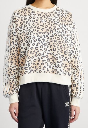 Kvinde iført en beige og sort leopardmønstret Adidas-sweatshirt med hvide striber på ærmerne og sorte Adidas-træningsbukser.