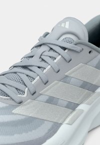 Chaussure de sport grise avec une tige en mesh texturé, trois bandes réfléchissantes, et une semelle amortie. Caractéristiques : forme arrondie et lacets sécurisés.