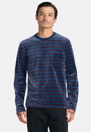 Homme portant une chemise bleue à manches longues avec de fines rayures horizontales rouges et une poche poitrine, debout devant un fond gris clair uni.