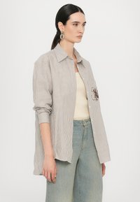 Camicia a righe in cotone marrone e bianco, con taschino sul petto con dettaglio ricamato, indossata sopra una canotta beige e pantaloni in denim.